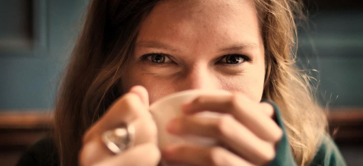 A close-up shot of a woman with long hair smiling slightly while holding a white mug up to her mouth, looking directly at the camera.