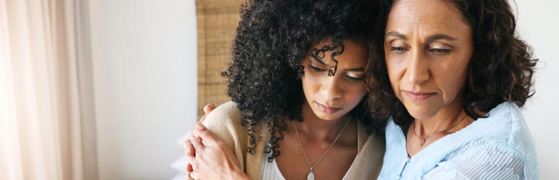 A close-up of two women of color looking somber and reflective. The woman on the right has her arm around the younger woman's shoulder in a supportive, comforting gesture.