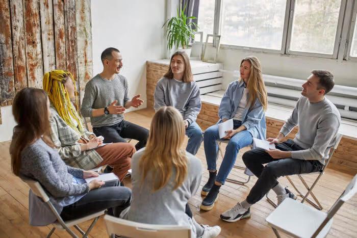 Six people, casually dressed, sit in a circle on folding chairs in a bright, rustic room with wooden floors and large windows, participating in a group therapy session or support meeting.