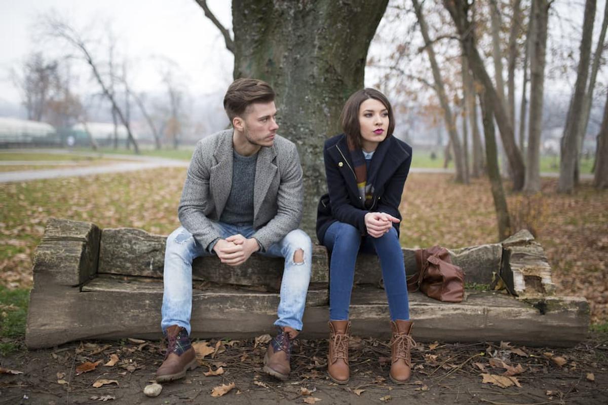 A young man and woman sit on a carved wooden log bench in a park, looking away from each other.