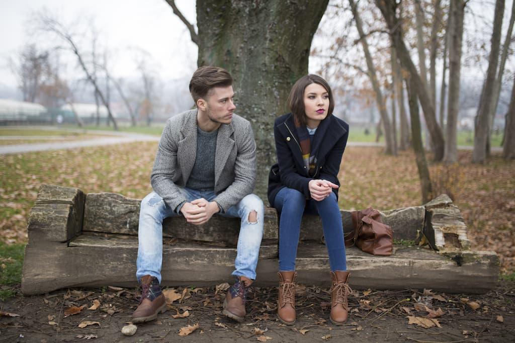 A young man and woman sit on a carved wooden log bench in a park, looking away from each other.
