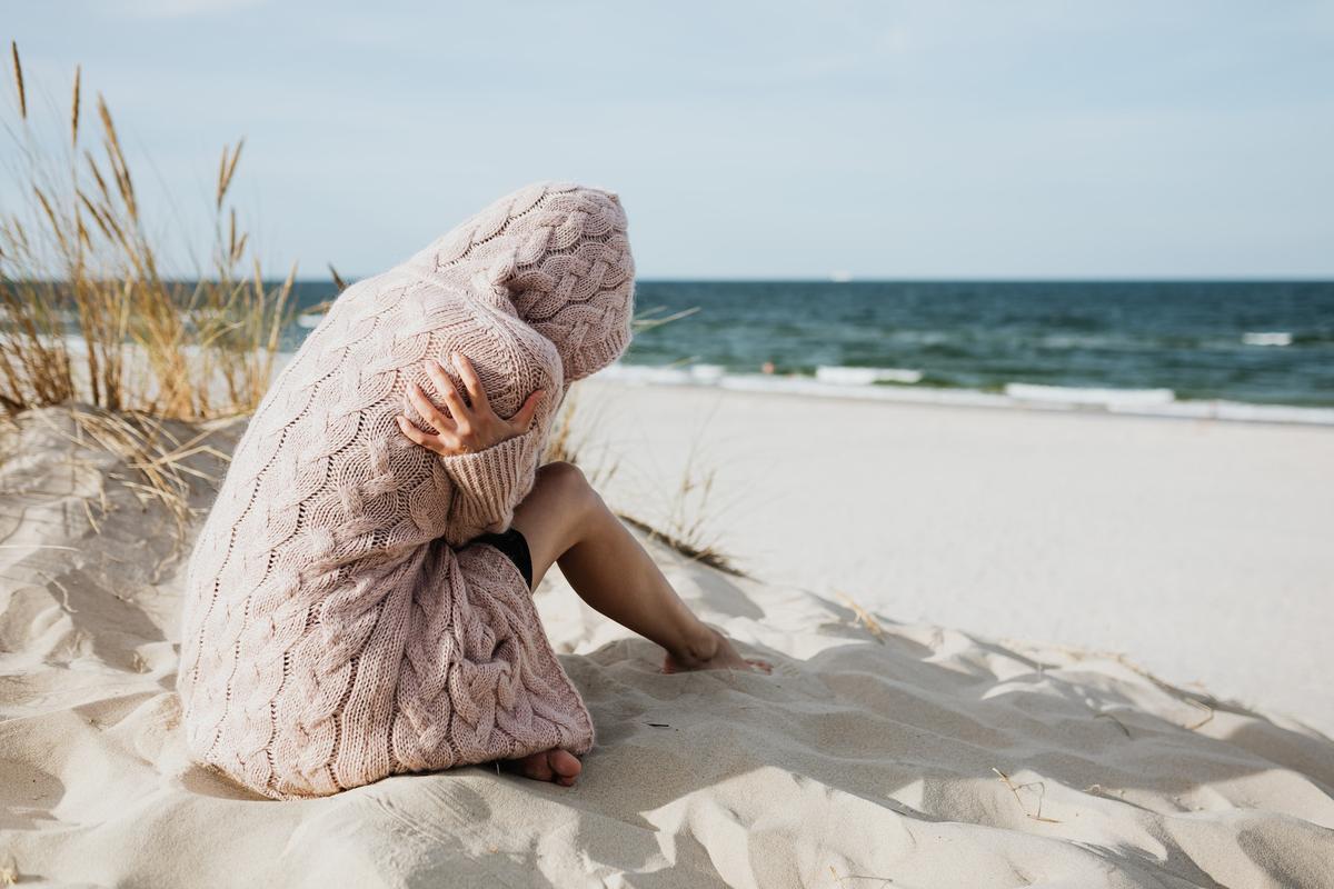A person sits curled up on a sandy beach dune, completely enveloped in a thick, oversized dusty-pink cable-knit sweater that covers their head.