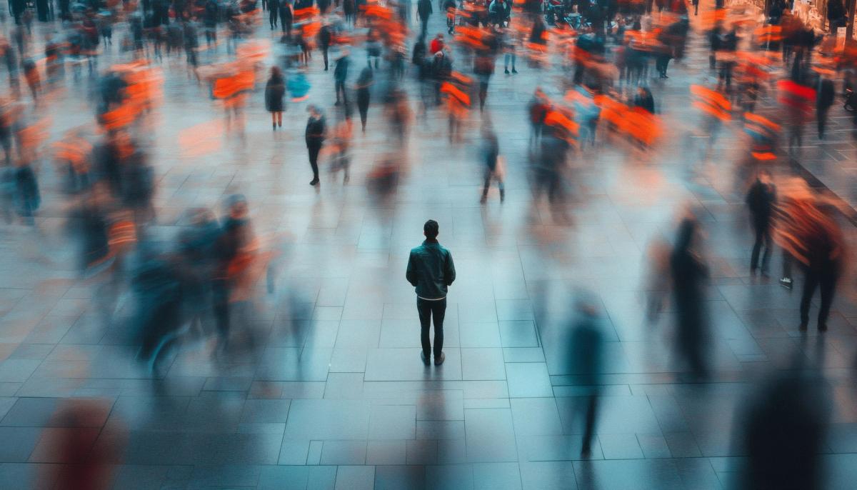 A lone man stands still in the center of a busy, blurred crowd of people in a large public space.
