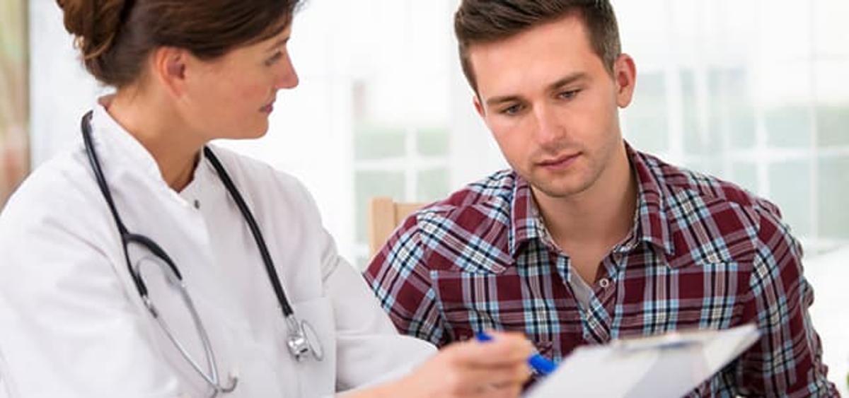 Female doctor discussing steroid addiction treatment with a young man during a clinic appointment, pointing at a form while he listens attentively in a bright exam room.