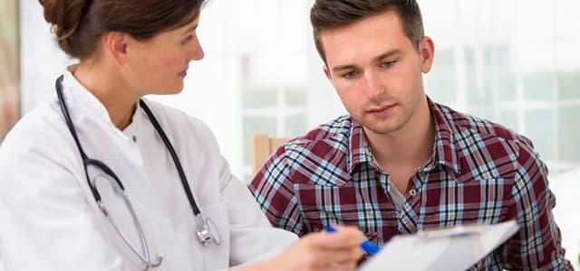Female doctor discussing steroid addiction treatment with a young man during a clinic appointment, pointing at a form while he listens attentively in a bright exam room.