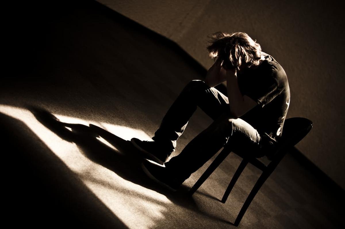 Depressed man sitting on a chair with head in hands in a dark room, symbolizing addiction, mental health struggles, anxiety, and the need for recovery treatment support.