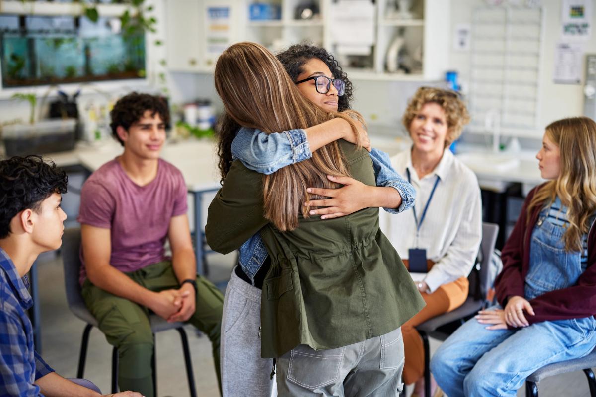 Teenage students sharing supportive hug during addiction recovery group session in safe school environment