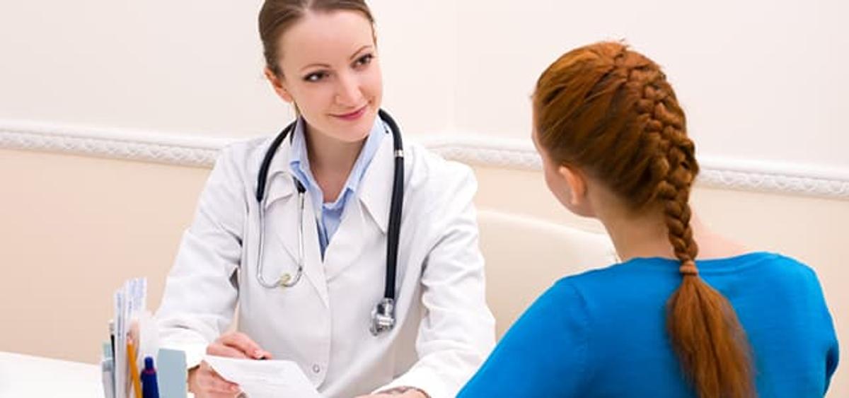 A smiling female doctor in a white coat and stethoscope around her neck sits at a desk, looking attentively at a female patient with red hair braided down her back who is facing away from the camera.