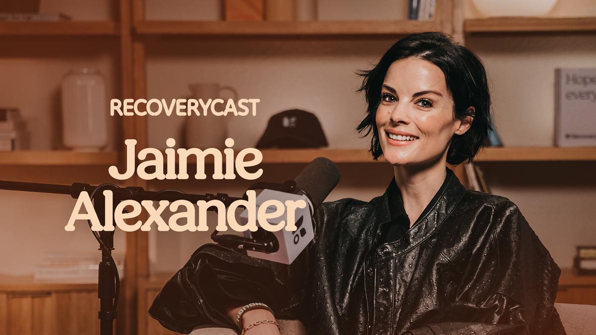 Actress Jaimie Alexander sits in a podcast studio smiling at the camera, wearing a black leather jacket and speaking into a microphone. The words “Recoverycast” and “Jaimie Alexander” appear on the left side of the image against a warm-toned background with wooden shelves.