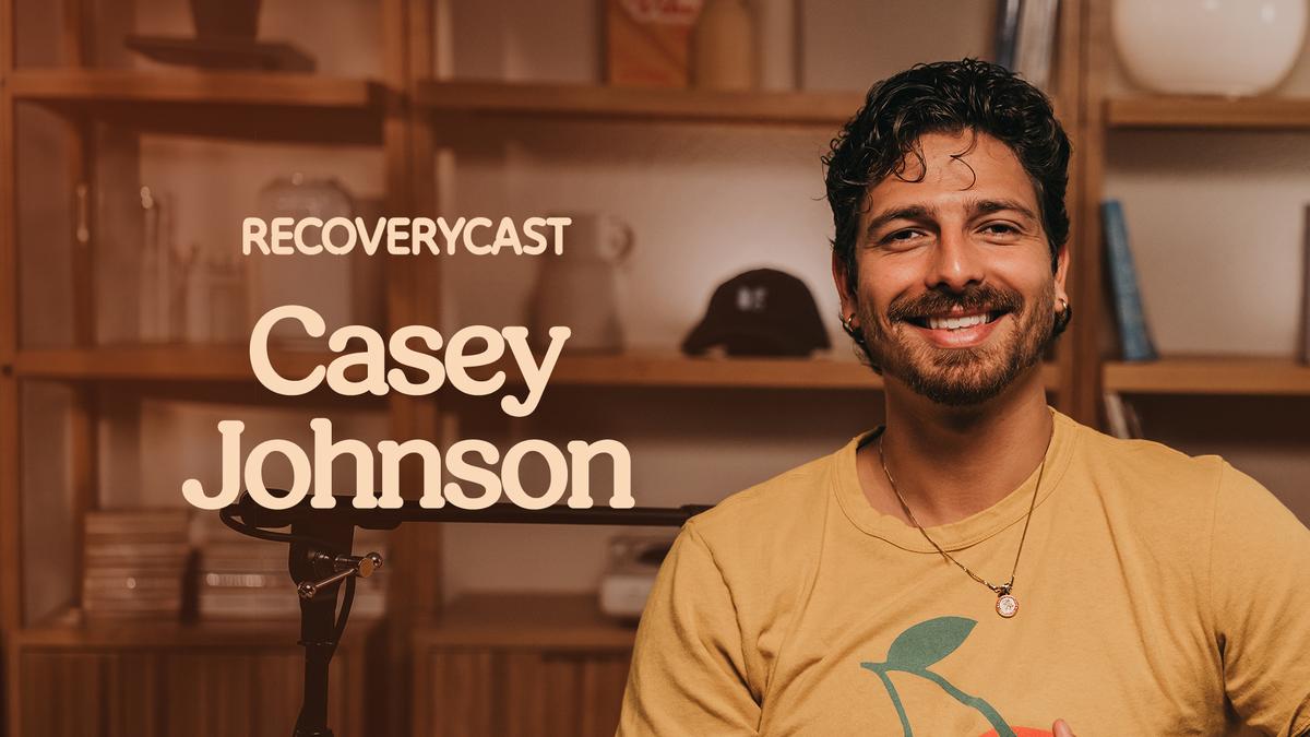 Recoverycast podcast episode featuring Casey Johnson smiling in a studio setting. Casey wears a yellow graphic T-shirt and sits beside a podcast microphone, with wooden shelves and decorative items in the background. The image includes the text "Recoverycast" and "Casey Johnson."