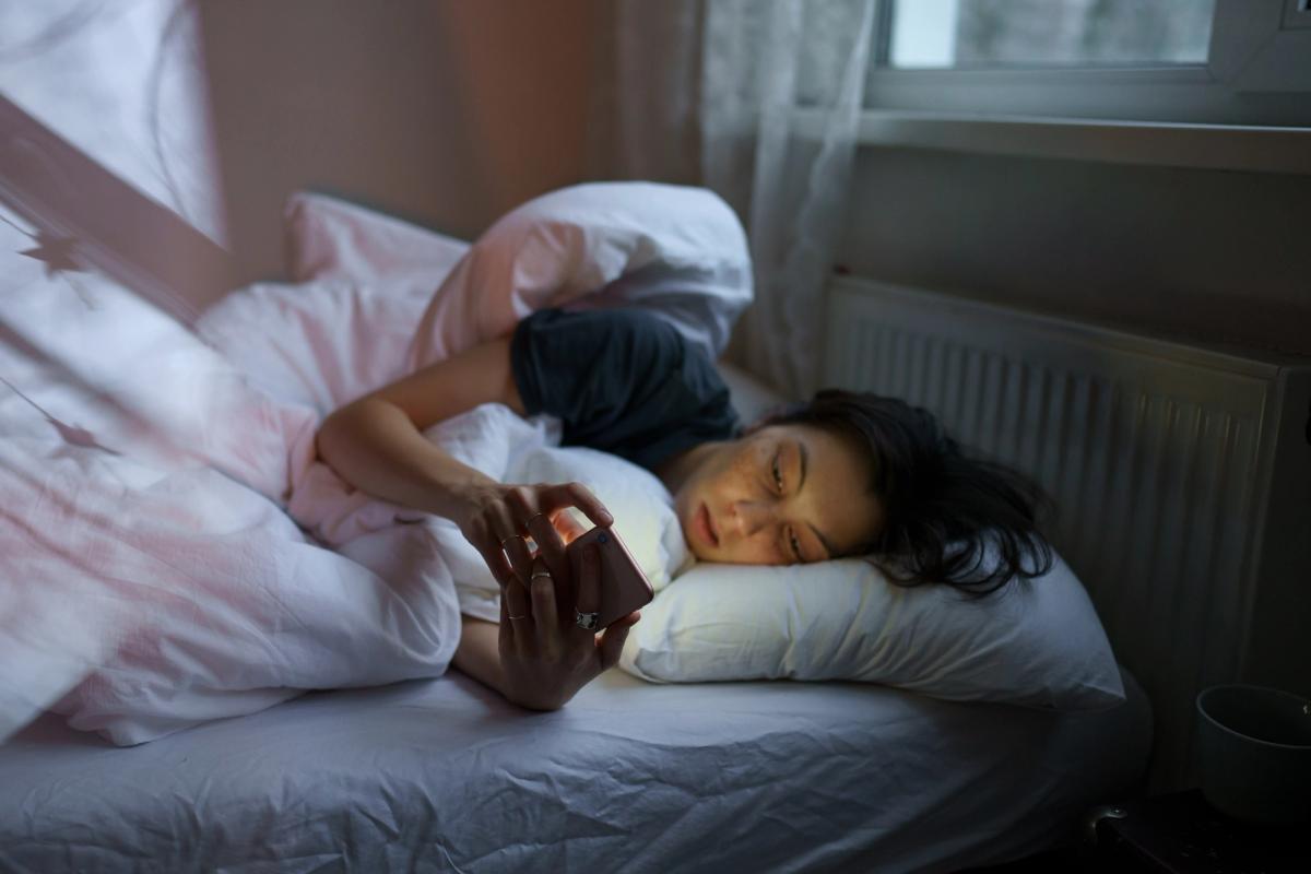 A young woman in bed, holding a smartphone with both hands while looking solemn.
