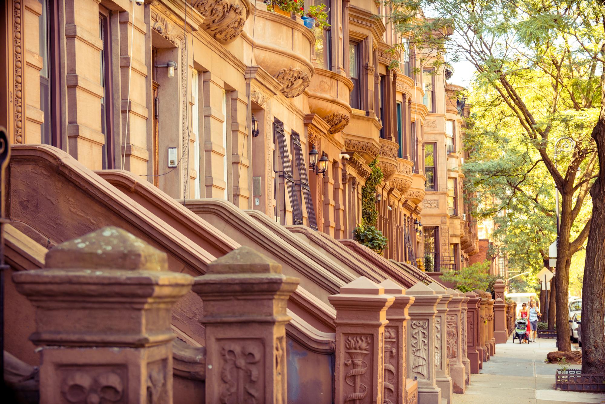 Row of classic brownstone townhouses with intricate stonework, bay windows, and prominent stoops lining a sunny, tree-lined street