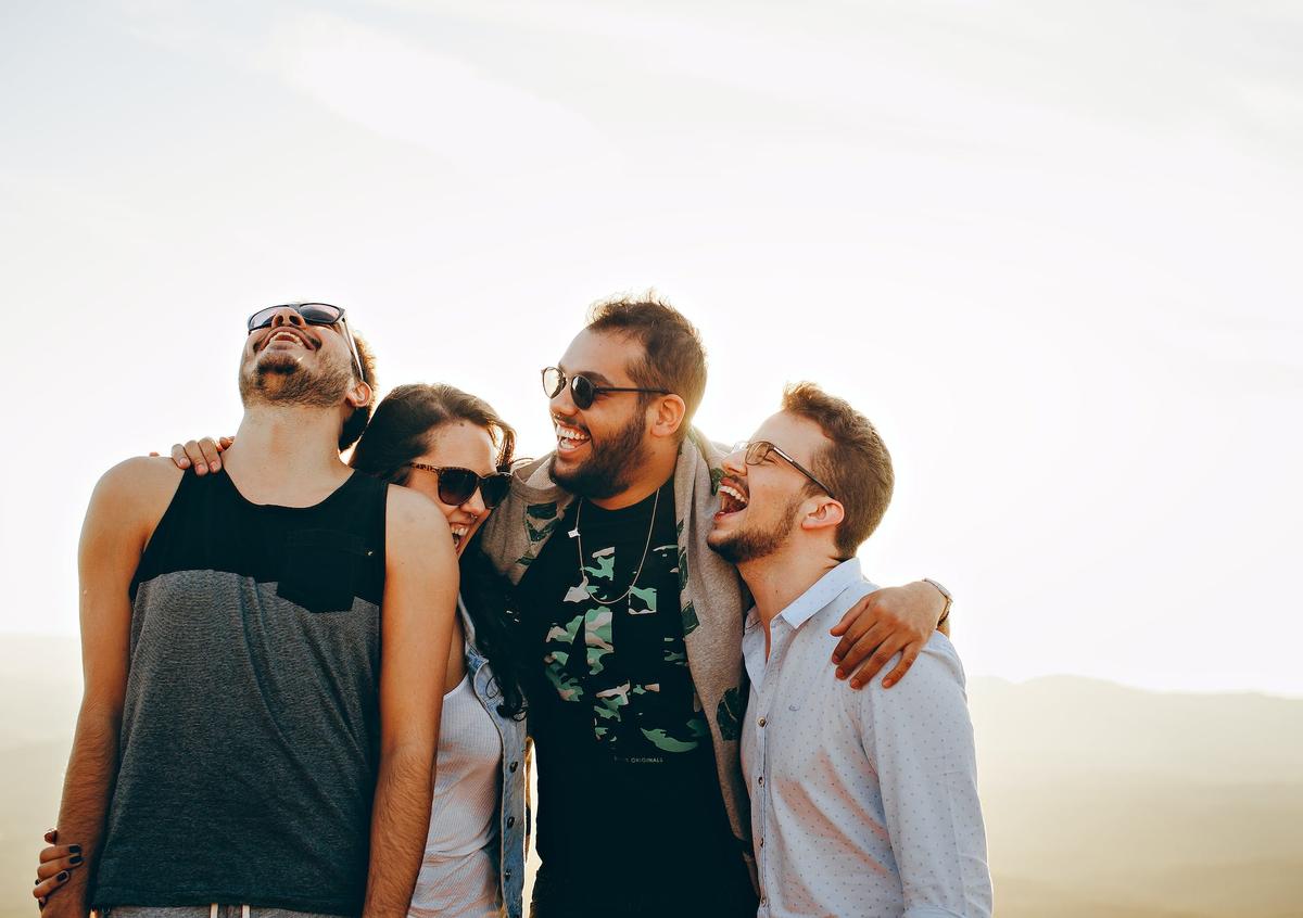 Four friends stand close together outdoors, arms around each other, laughing and smiling in the bright sunlight with a hazy landscape behind them.