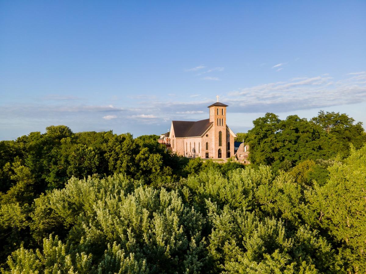 An aerial view of a stone church with a tall bell tower rising above a lush, green forest under a clear blue sky.
