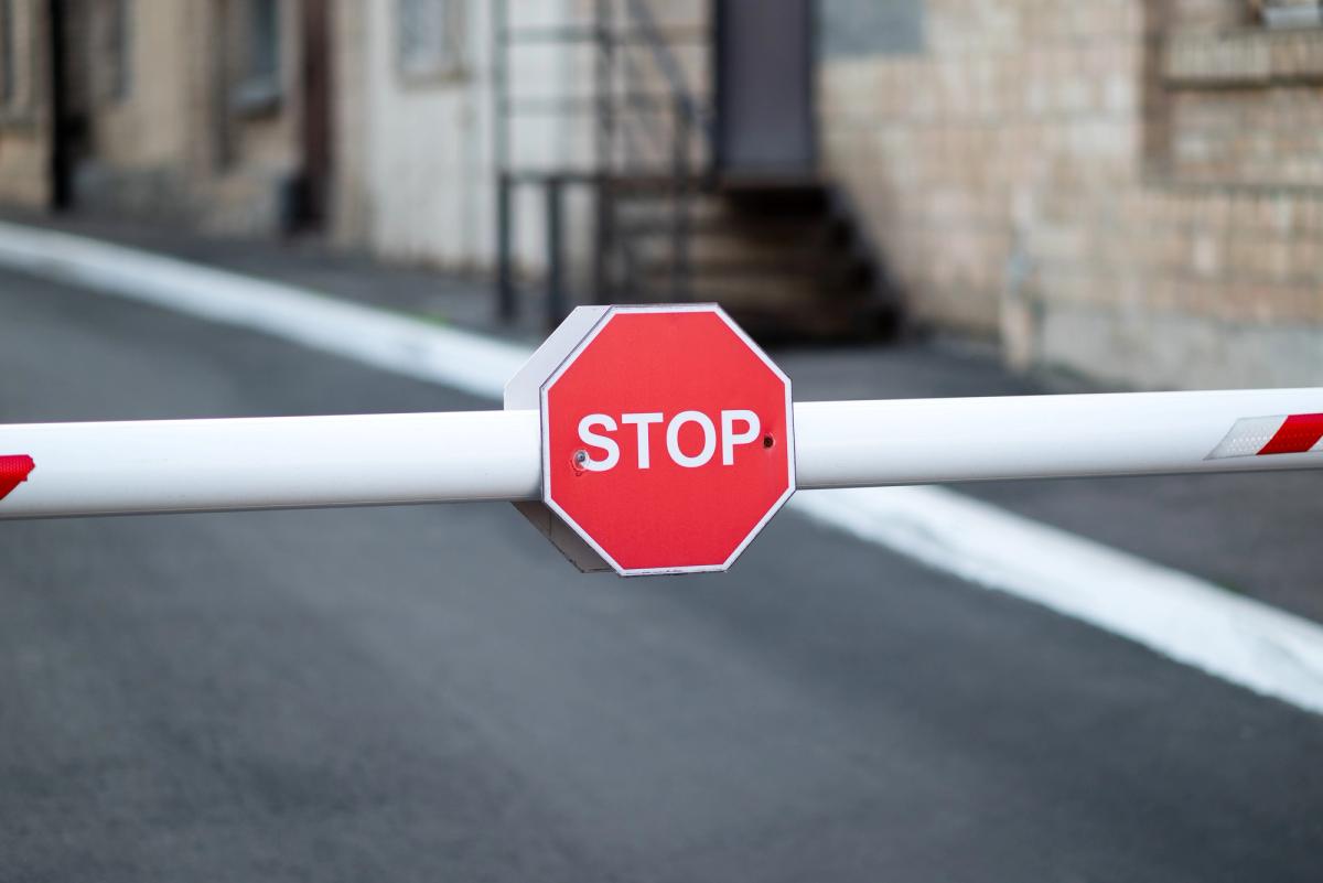 A close-up of a white automatic parking arm barrier in the down position, featuring a small red octagonal sign in the center with the word "STOP" in white capital letters.