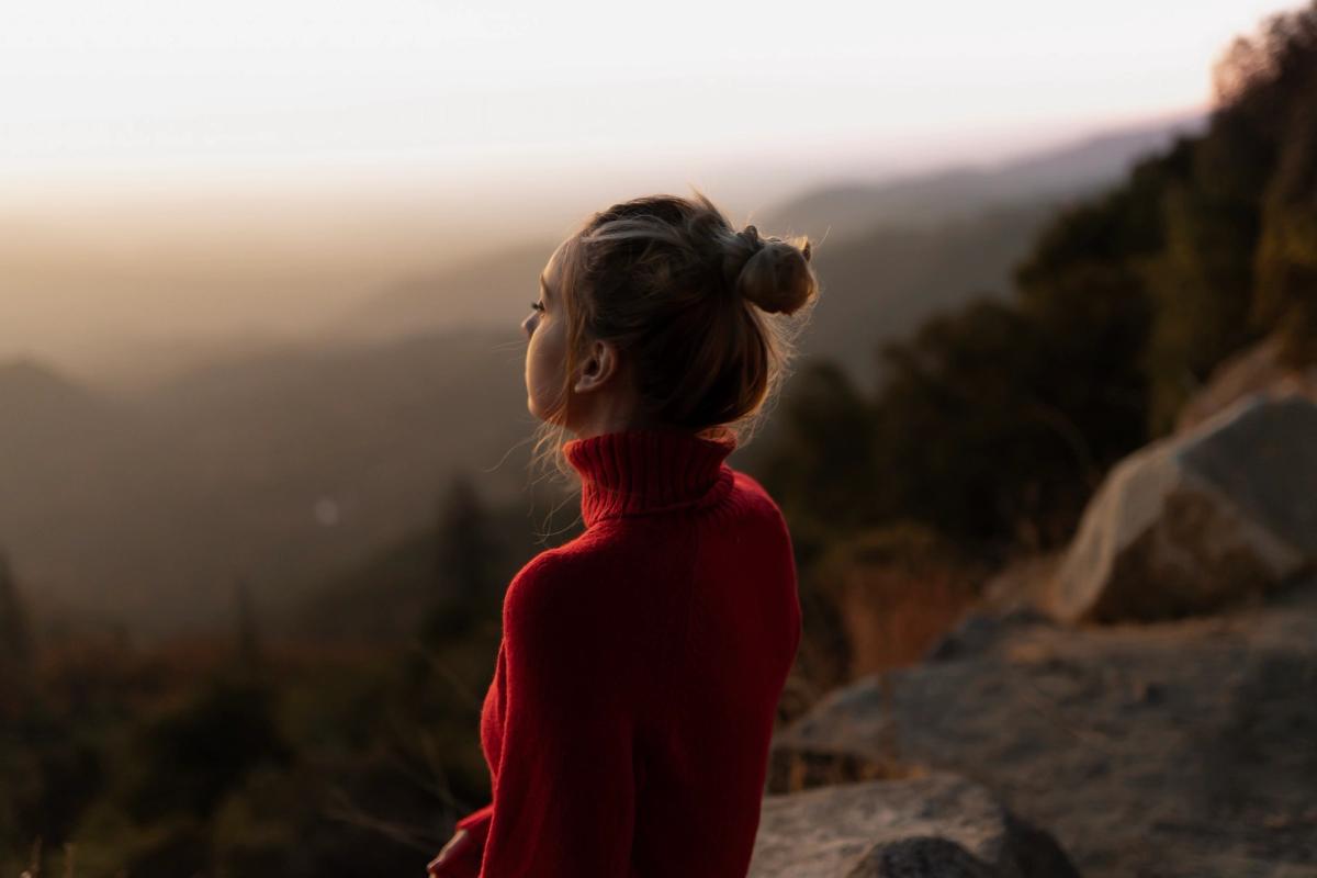 Woman in a red sweater standing on a mountain overlook at sunset, reflecting thoughtfully on whether she may have a drug problem.