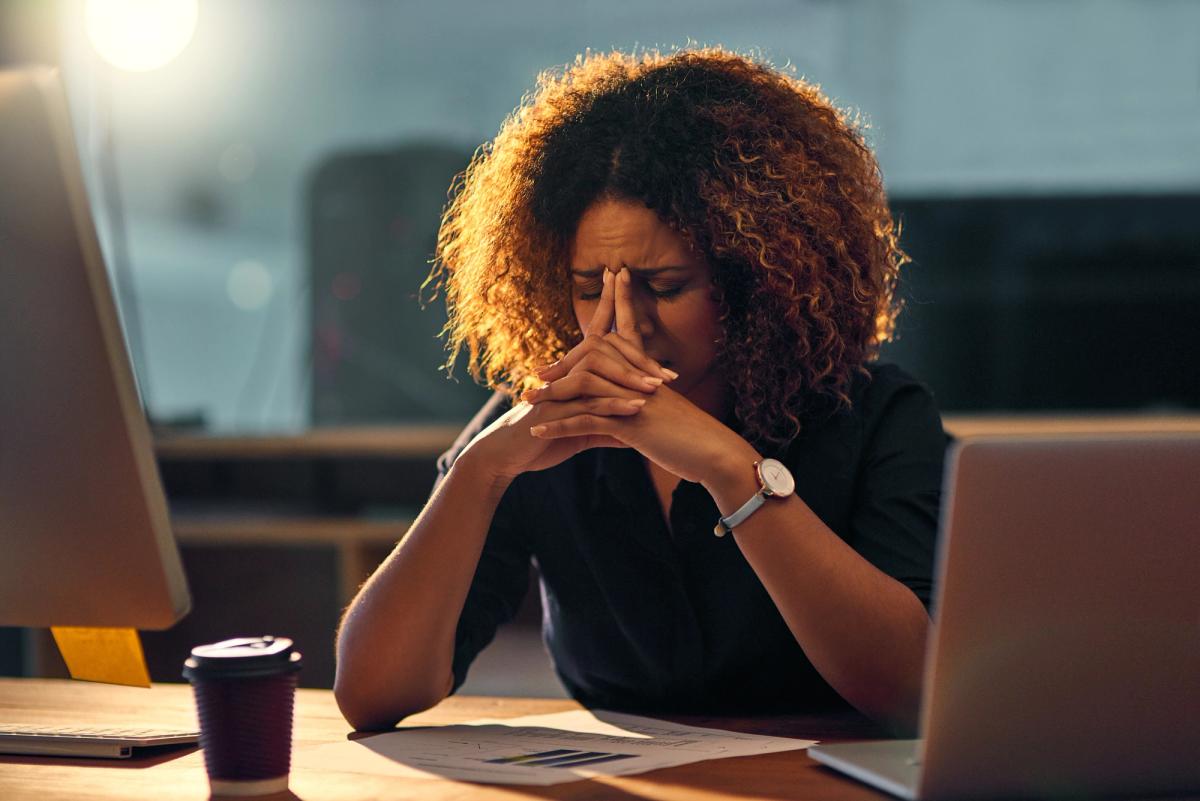 Woman sitting at a desk with her hands clasped to her face, appearing stressed while looking down at paperwork between two laptops.