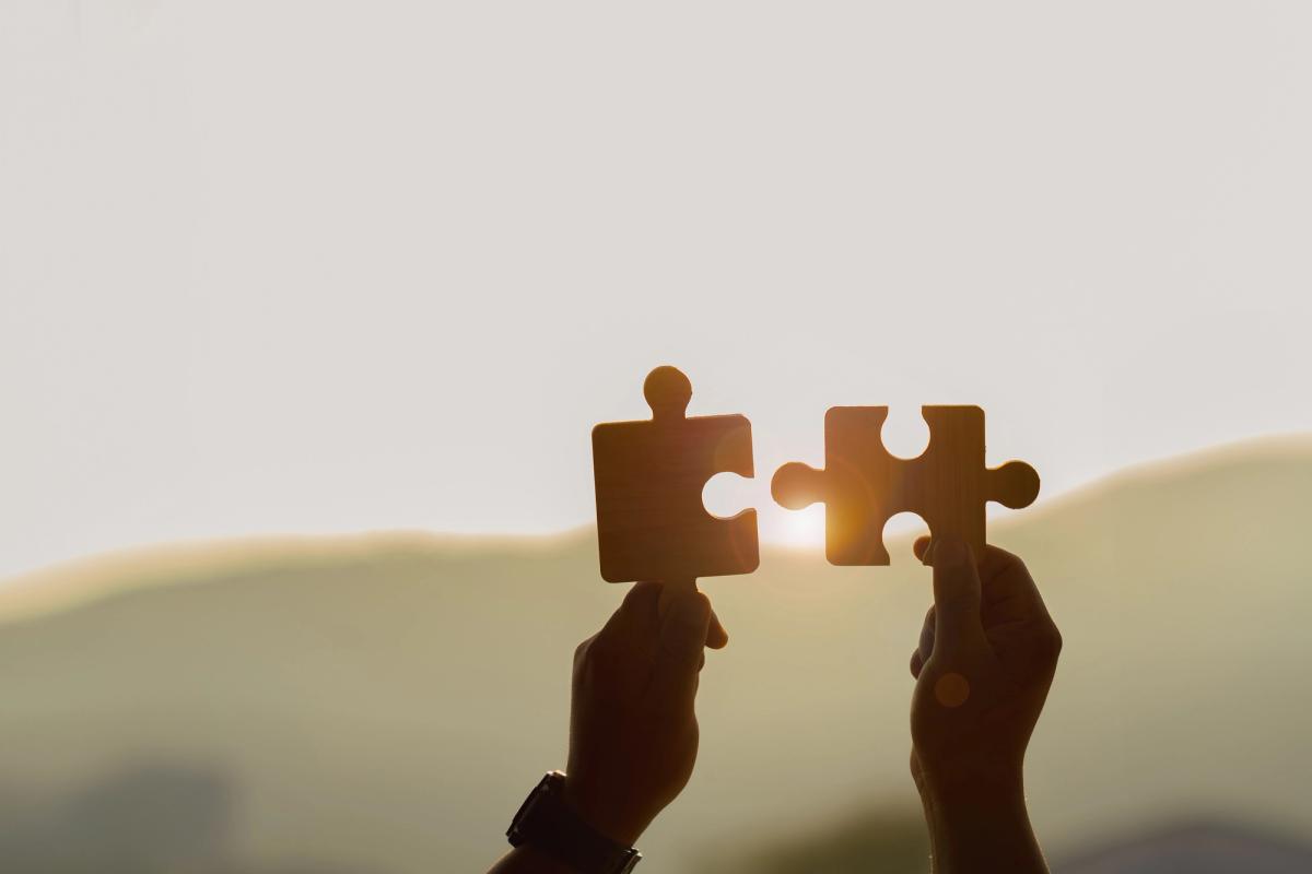 Two hands hold up two large puzzle pieces against a sunset background, positioned as if they are about to be joined together.