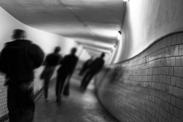 A black and white, motion-blurred long exposure photograph of several blurred human figures walking quickly down a dimly lit, curving pedestrian tunnel with tiled walls.