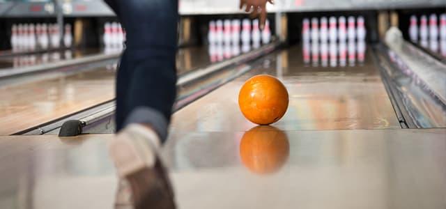 An orange bowling ball rolls down a wooden lane toward white pins.