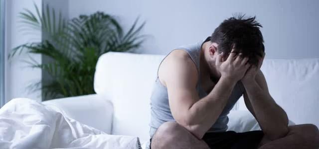 A distressed man in a tank top sits on a white couch, leaning forward with his head in his hands, suggesting stress, anxiety, or sadness.