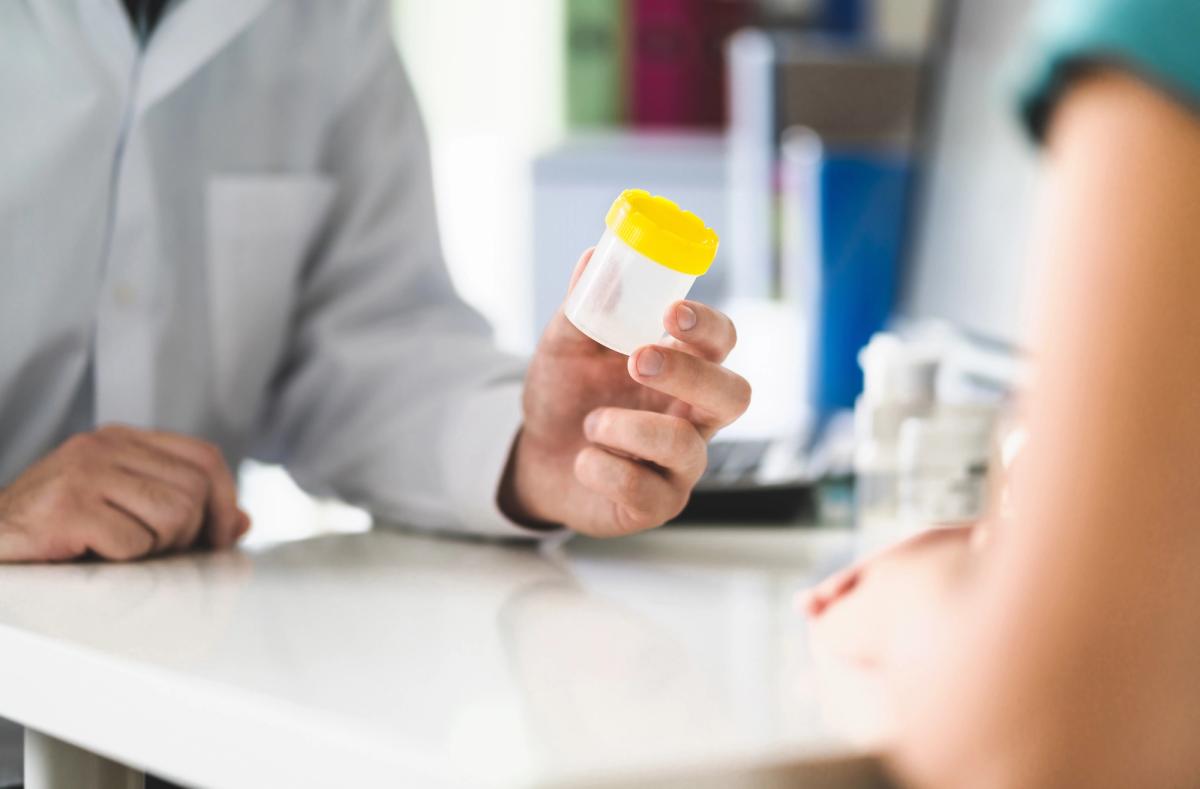 A healthcare provider in a white coat holds up a clear specimen cup with a yellow lid while speaking with a patient across a desk.