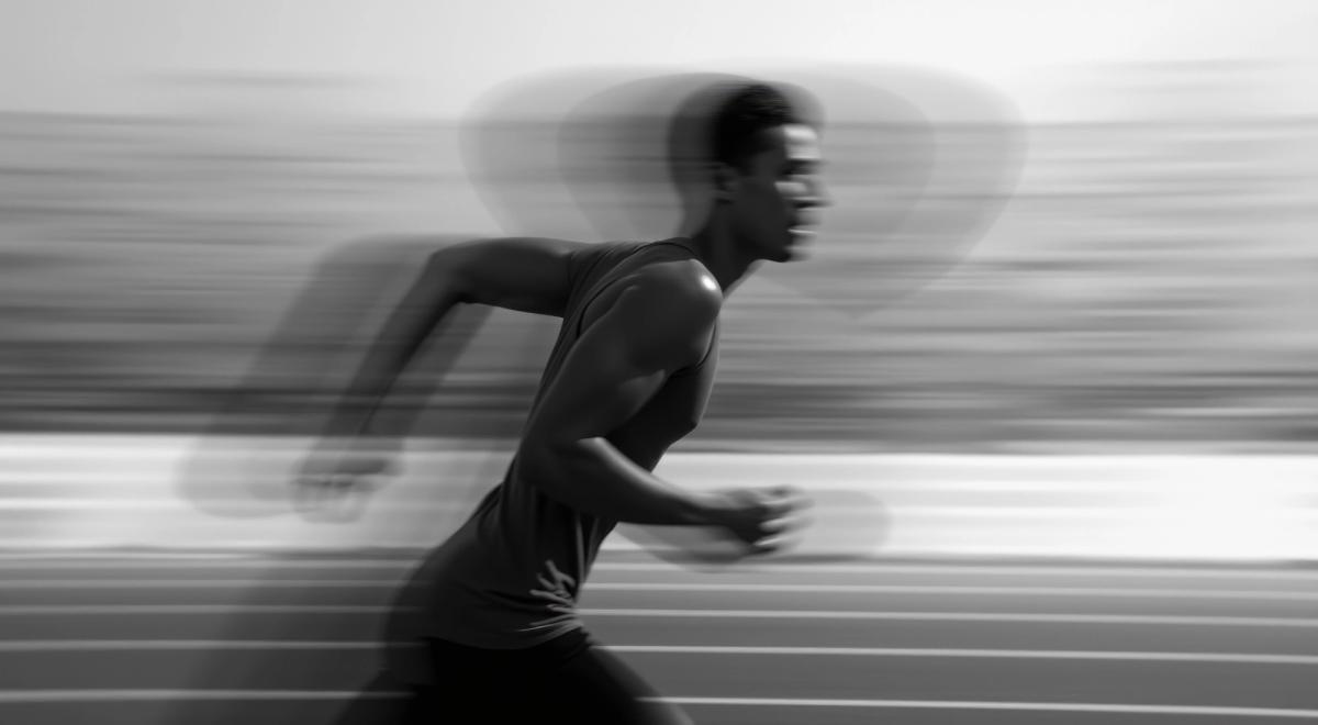 A black and white, motion-blurred photograph of a male athlete sprinting on a track.