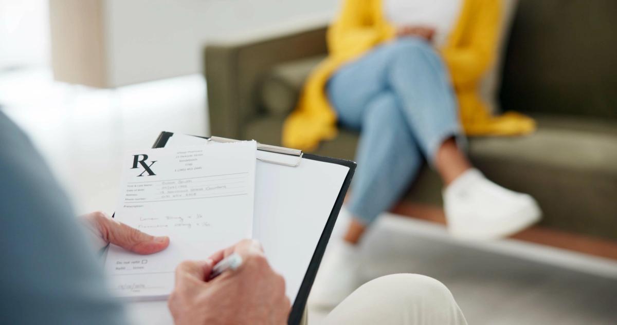 Close-up of a clinician holding a clipboard with a prescription form while a patient sits blurred in the background on a couch, wearing a yellow cardigan and jeans.