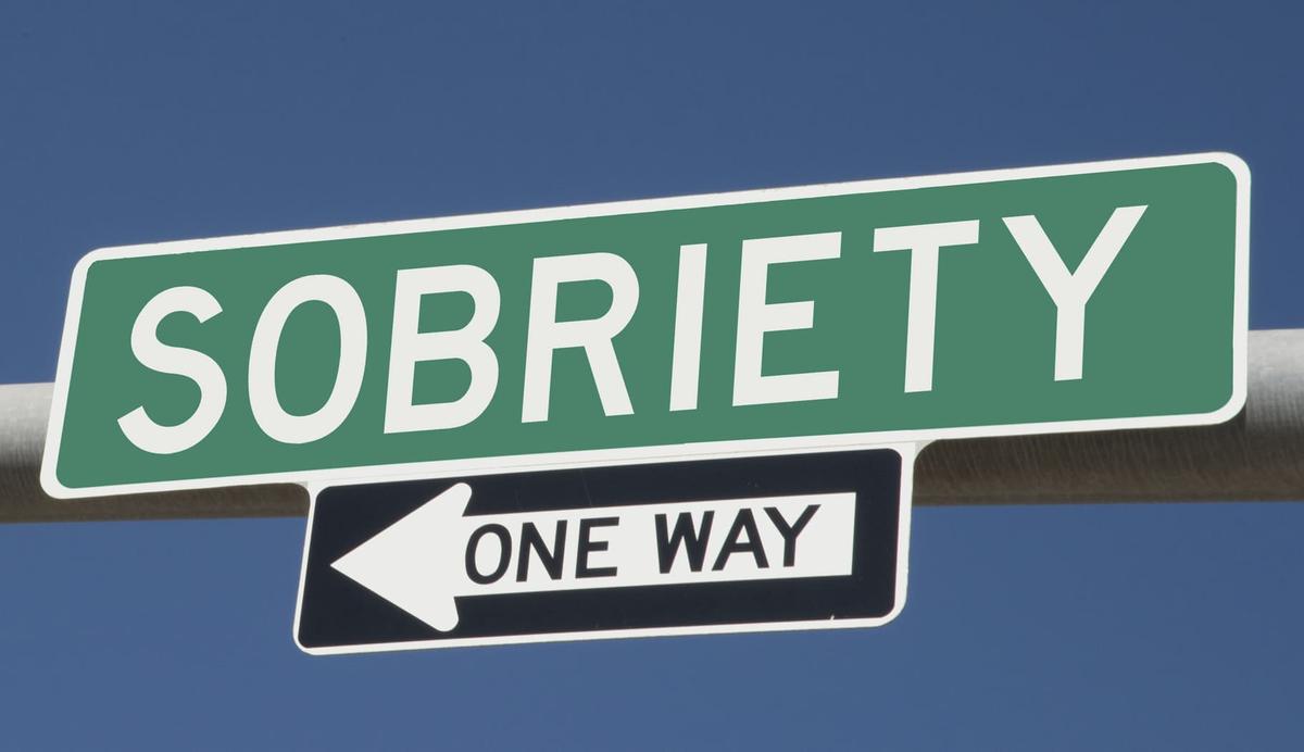 A green street sign with the word "SOBRIETY" in white capital letters. Below it, a black and white "ONE WAY" sign points to the left against a clear blue sky