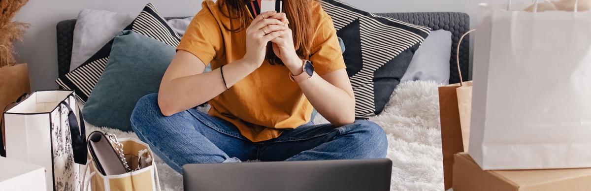Woman sitting cross-legged on a couch holding a credit card in front of a laptop, surrounded by shopping bags and packages, illustrating shopping addiction and compulsive online spending.