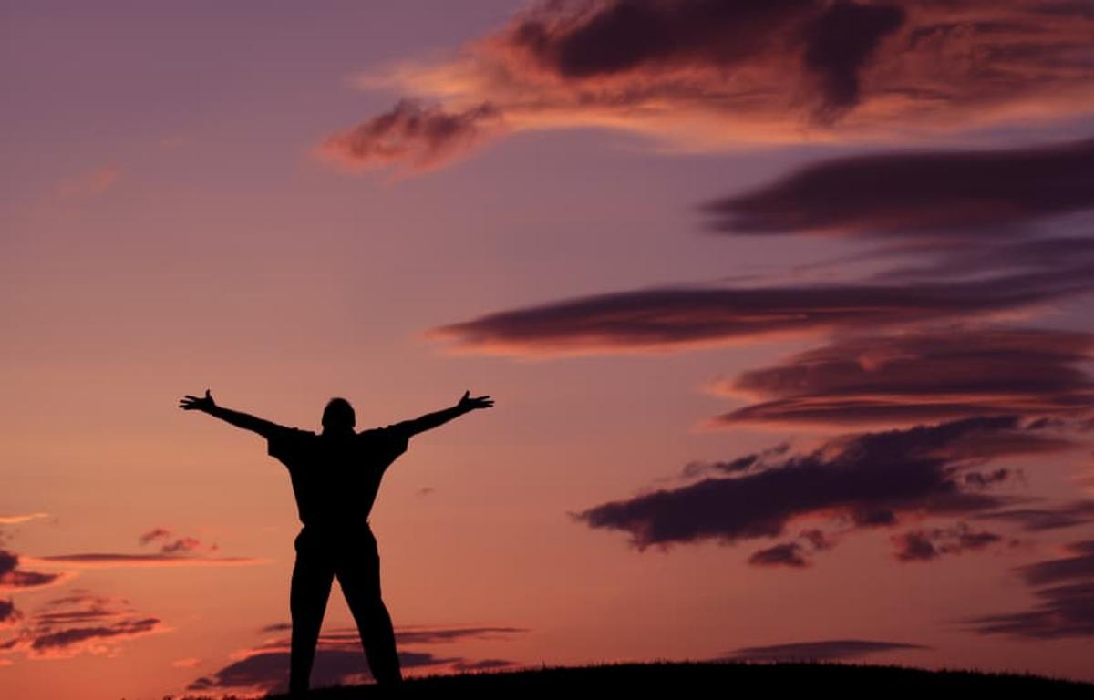 A silhouette of a person standing on a hilltop with their arms spread wide against a vibrant purple and orange sunset sky.
