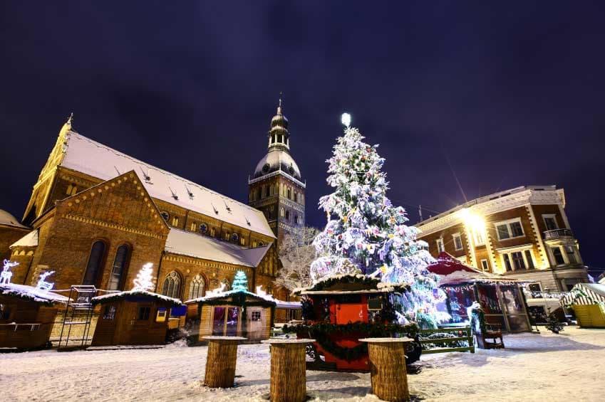 A snowy Christmas market at night in front of a large, illuminated cathedral and a lit clock tower.