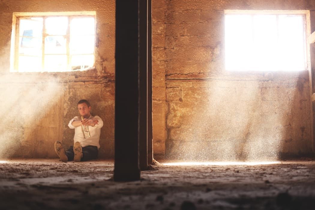 A man sits on the floor of a dusty, rustic room with sunbeams streaming through two windows.