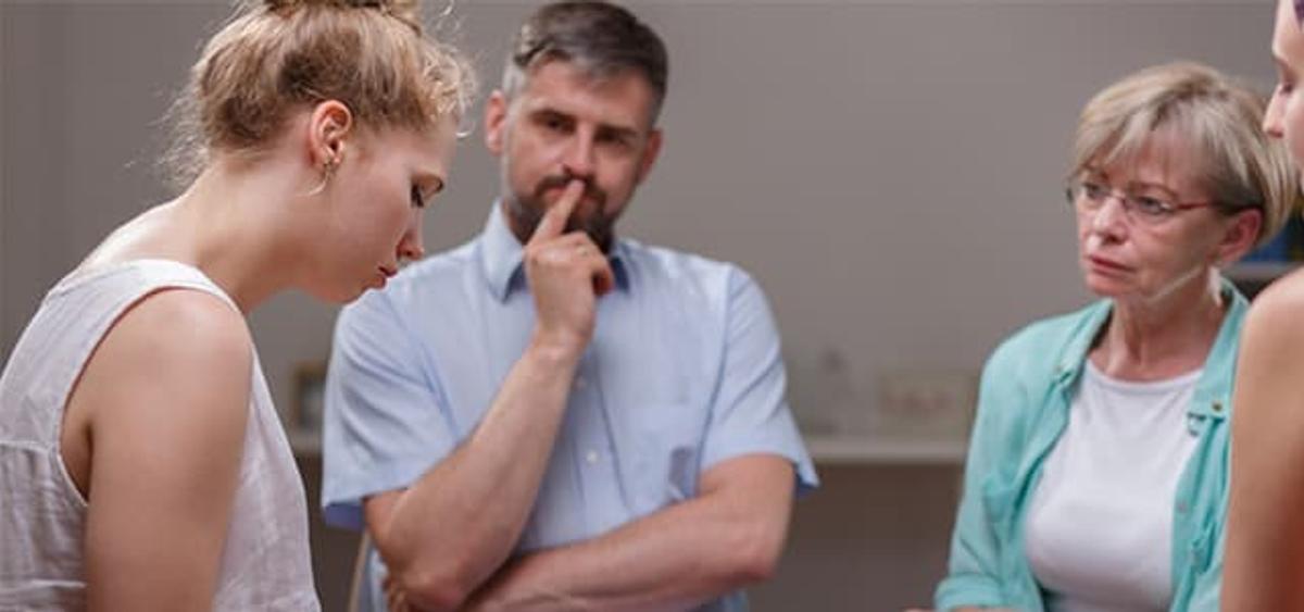 Small group conversation resembling an Alcoholics Anonymous meeting, with a young woman looking down thoughtfully while two older adults listen attentively, one man with his hand on his chin and a woman watching with concern, suggesting support and reflection in recovery.