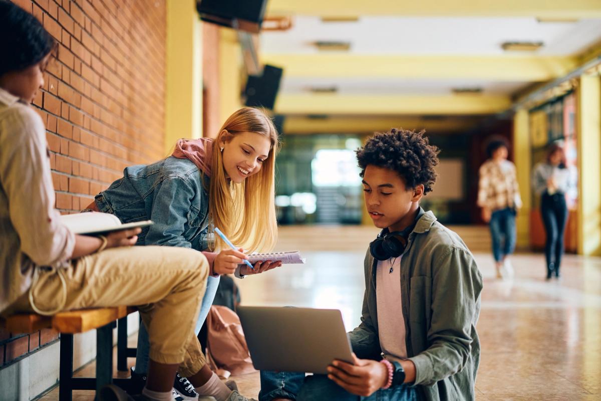 Group of teenagers sitting in a school hallway, collaborating over a laptop and notebook, with one student wearing headphones around their neck while others look on.