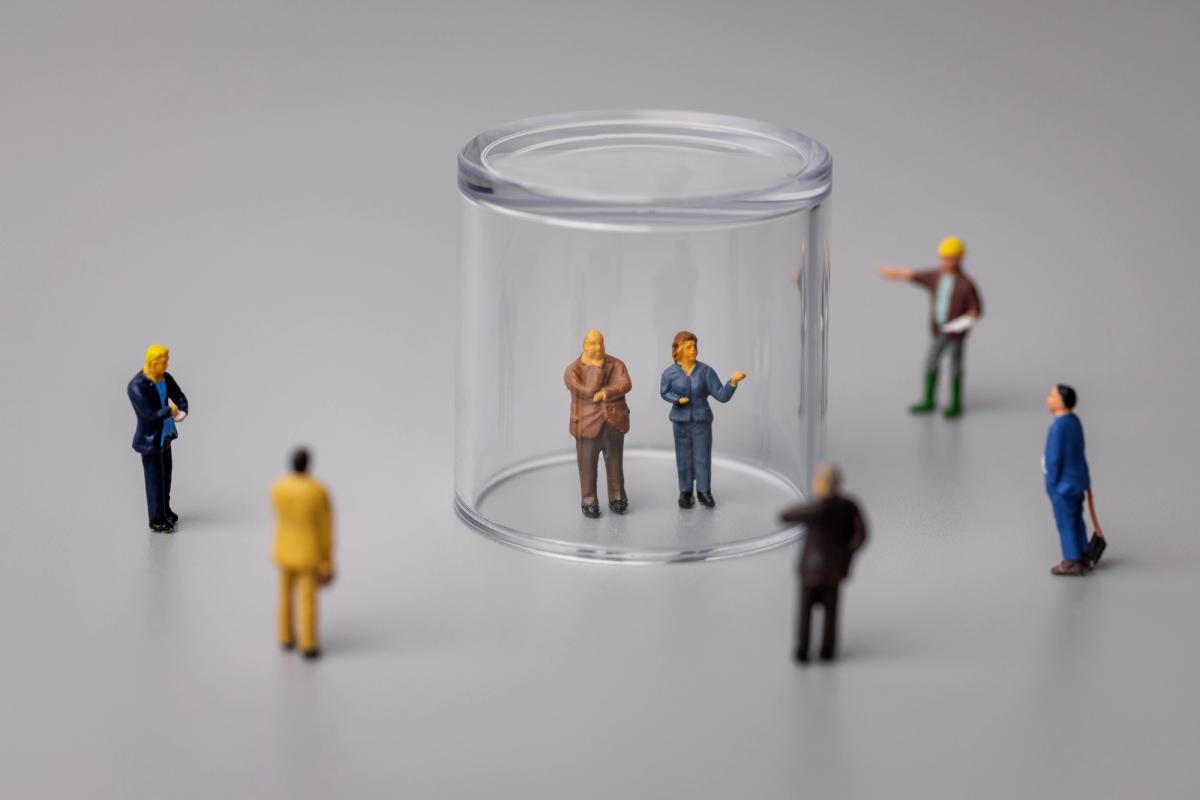 Miniature figures surround two people standing under a clear plastic dome, symbolizing social isolation, mental health stigma, and the need for support and recovery treatment.