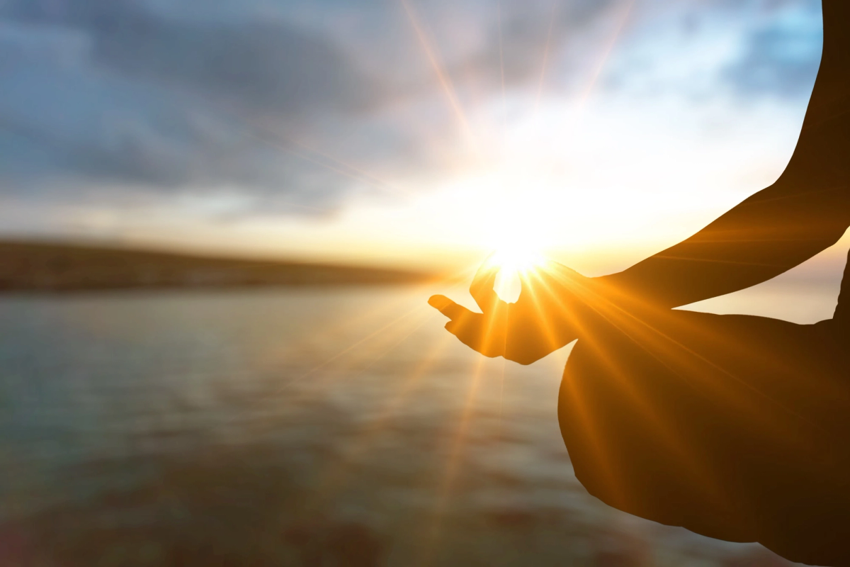 Person meditating in lotus position by the water at sunrise with sun rays shining through hand gesture, symbolizing peace, mindfulness, and inner balance.