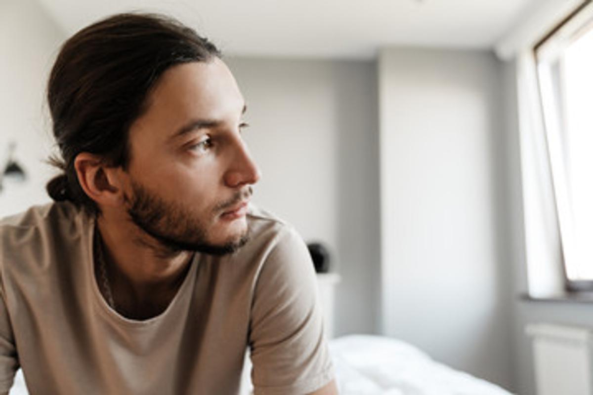 Close-up of a contemplative young man with long dark hair tied back, wearing a tan t-shirt and looking off to the right, sitting in a bright, modern bedroom.