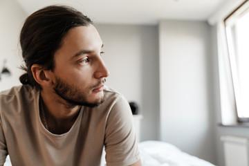 Close-up of a contemplative young man with long dark hair tied back, wearing a tan t-shirt and looking off to the right, sitting in a bright, modern bedroom.