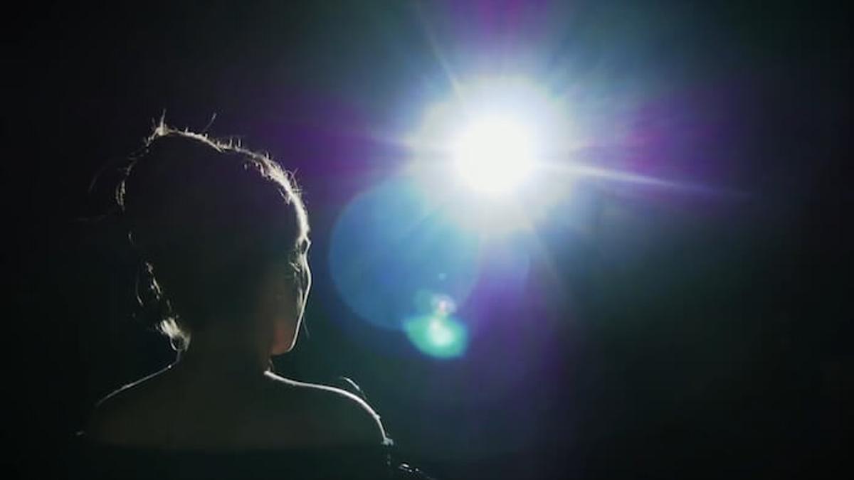 Silhouette of a person with their hair in a bun looking toward a bright, glowing light in a dark room.