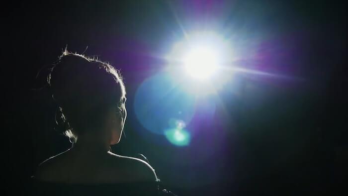 Silhouette of a person with their hair in a bun looking toward a bright, glowing light in a dark room.