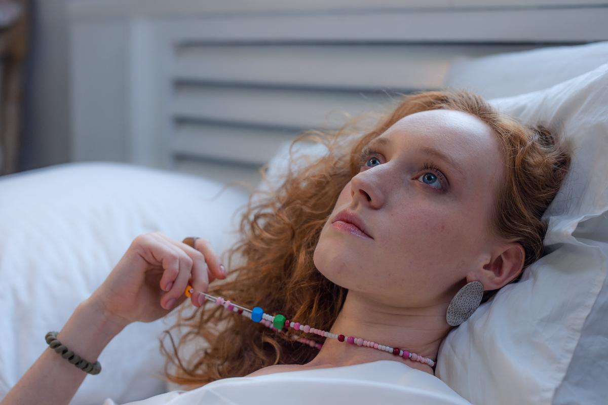 A young woman with red curly hair lying on white pillows, looking upward with a thoughtful expression.