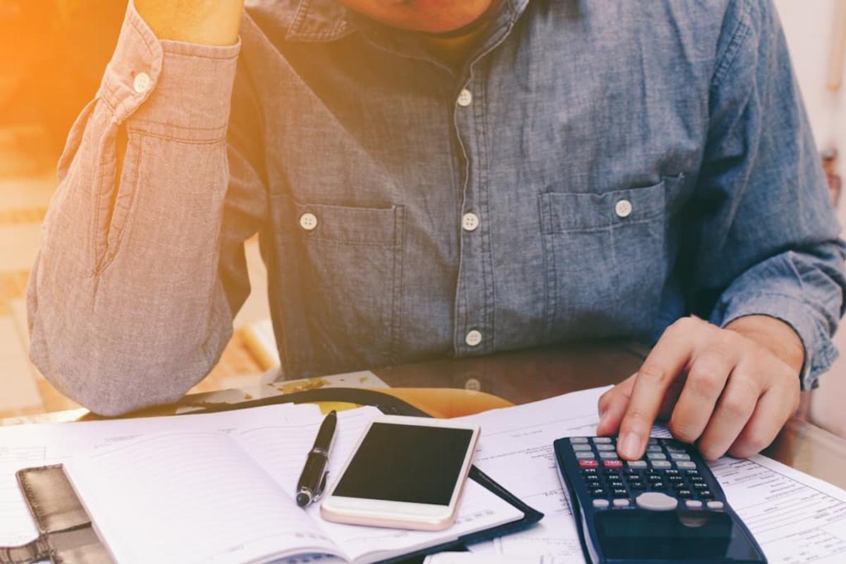 A person in a blue button-down shirt sitting at a desk, using a calculator to review financial documents, a notebook, and a smartphone.