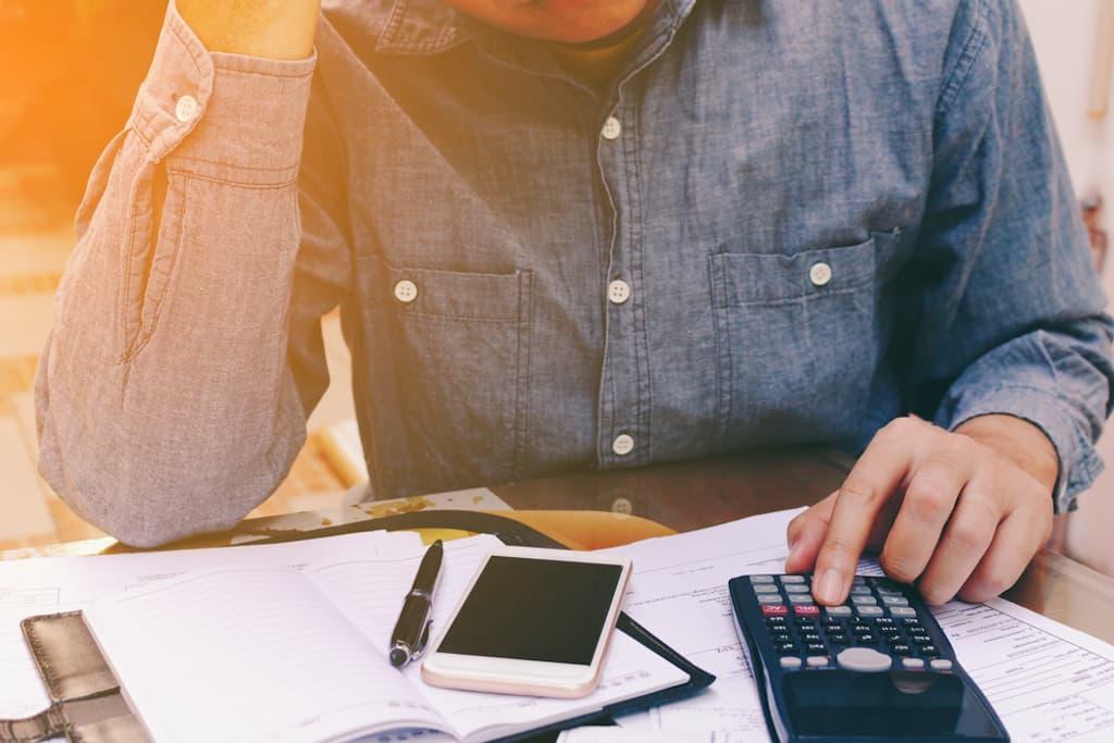 A person in a blue button-down shirt sitting at a desk, using a calculator to review financial documents, a notebook, and a smartphone.
