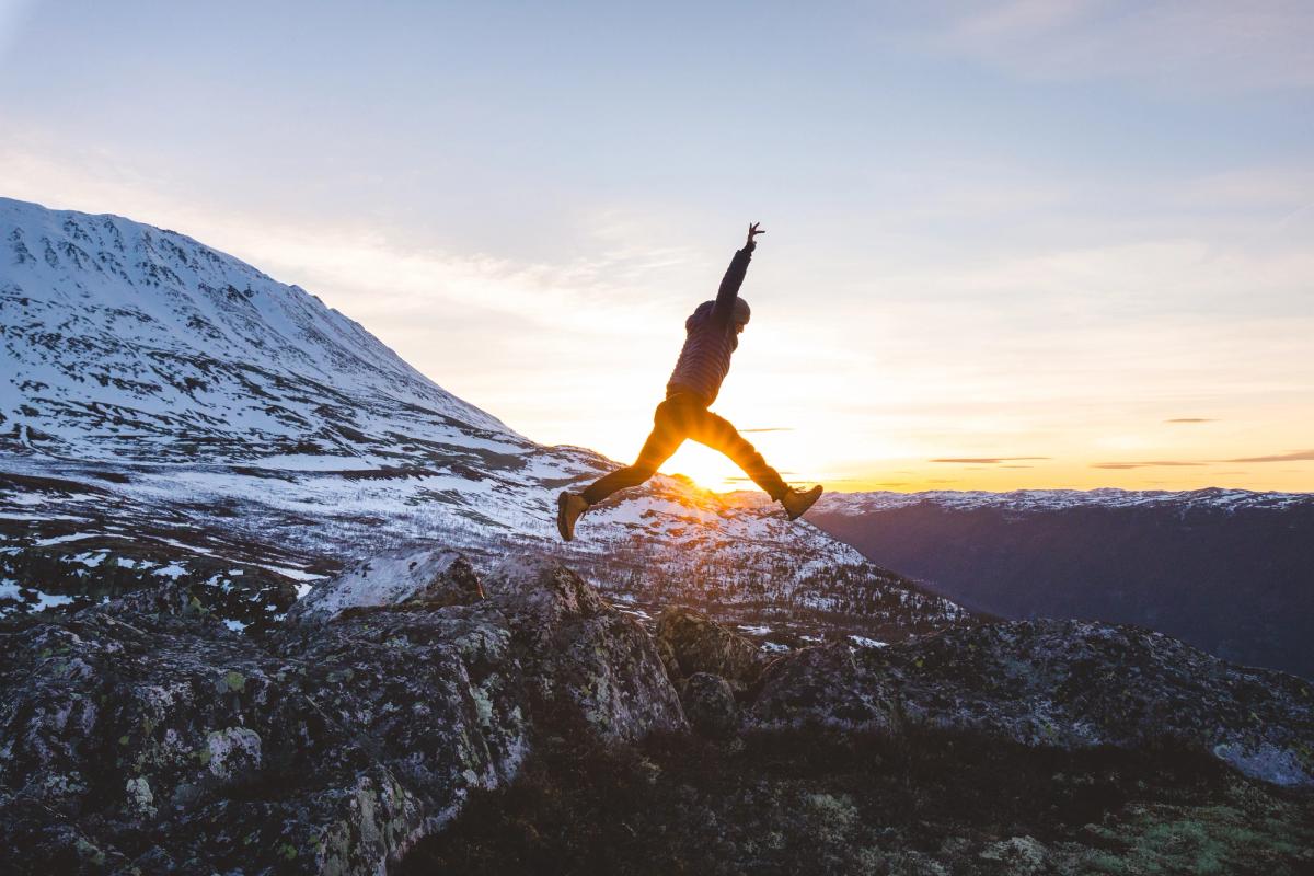 Person jumping between rocks at sunset with snowy mountains in the background.