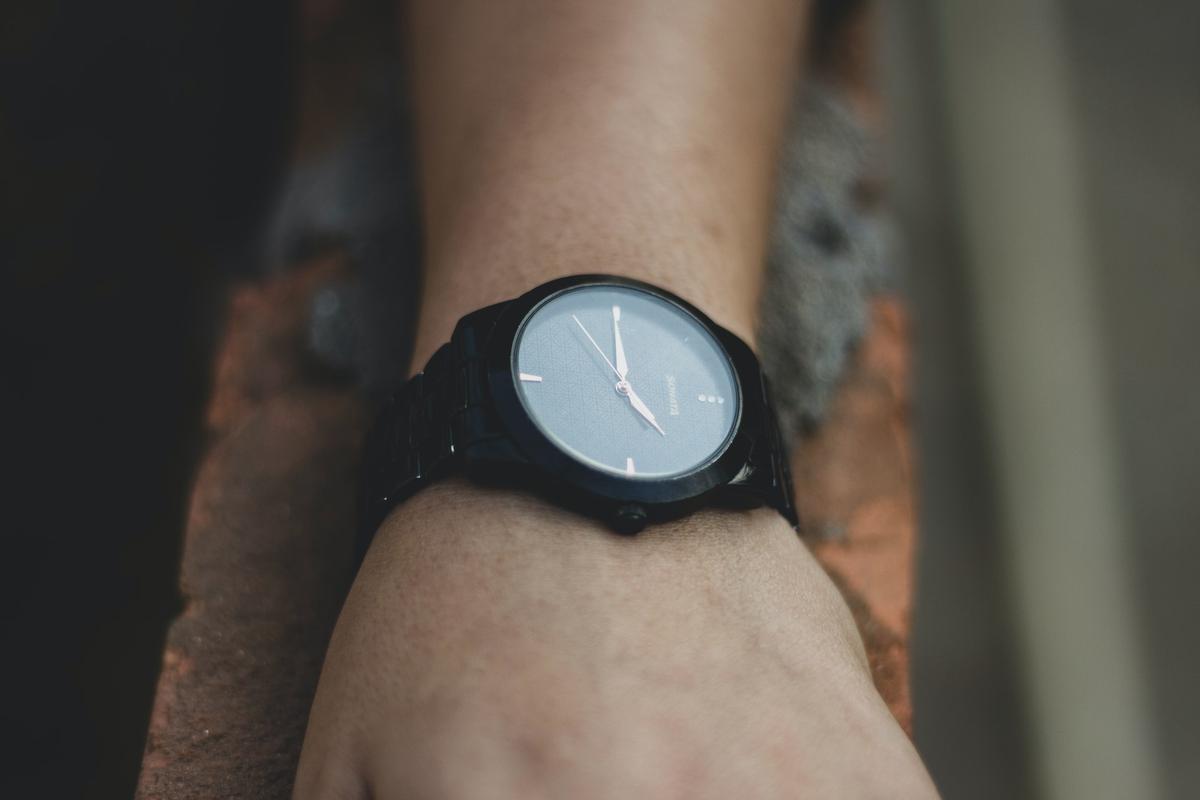 Close-up, high-angle shot of a person's wrist wearing a minimalist black analog watch with a dark face and copper-colored hands, resting against a weathered red brick.