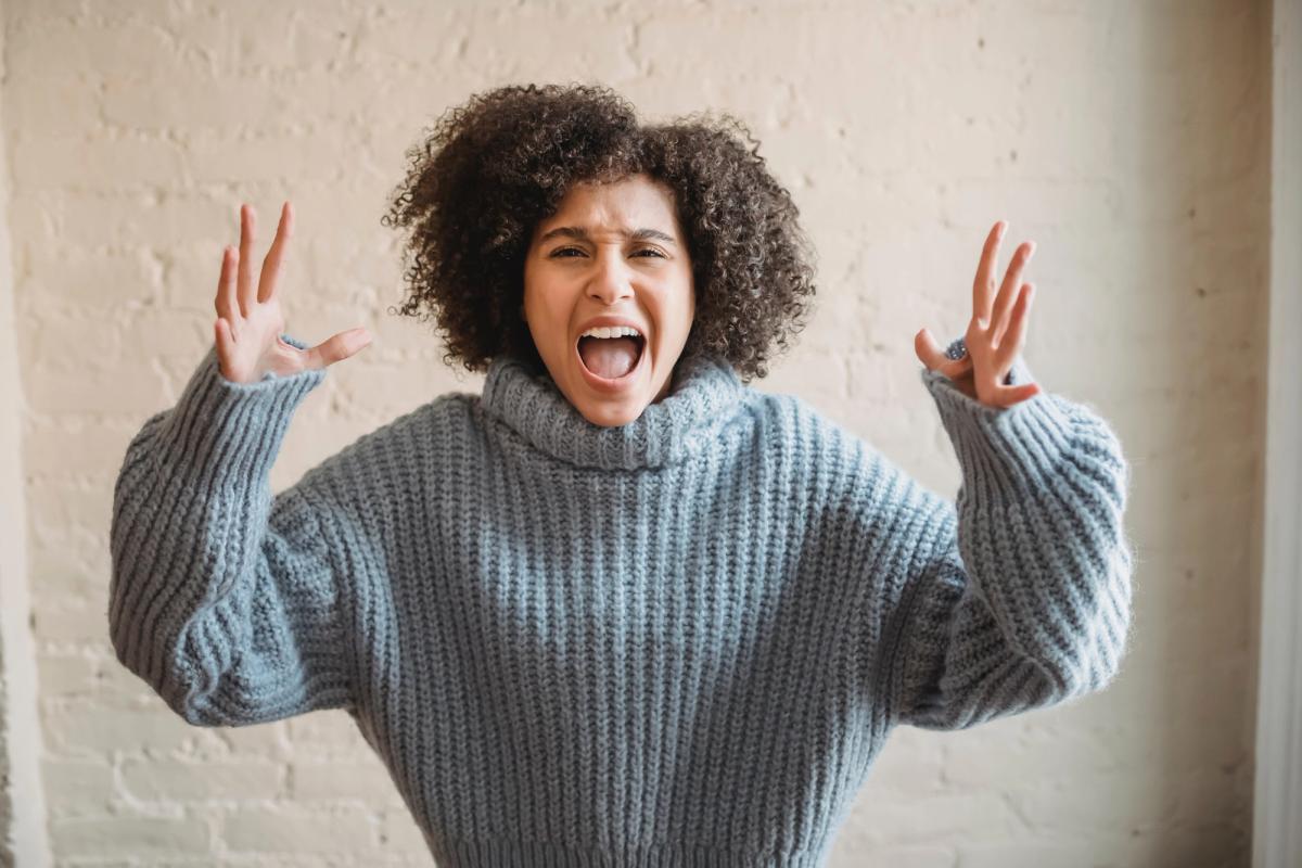 A woman with curly hair wearing a blue turtleneck sweater shouting with her hands raised.