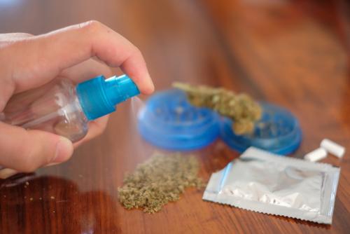 A hand spraying a clear liquid from a small spray bottle onto a pile of ground cannabis on a wooden surface.