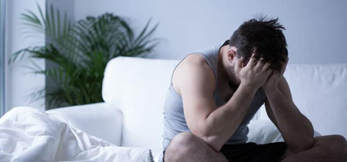 A man sits hunched on the edge of a bed, head in his hands, with rumpled white sheets and a houseplant in the background—suggesting distress or opioid withdrawal symptoms.