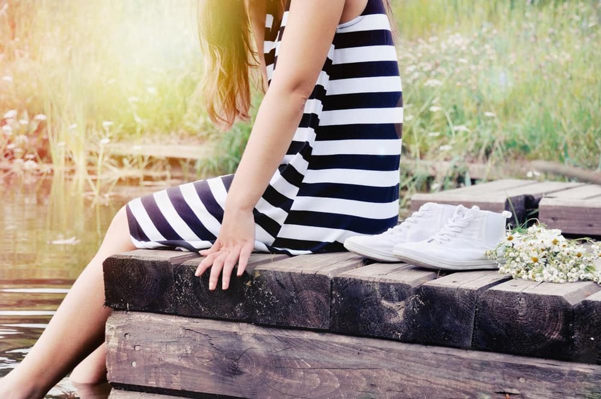 Woman in a black-and-white striped dress sitting on a wooden dock by the lake, barefoot with white sneakers and flowers beside her in warm sunlight.